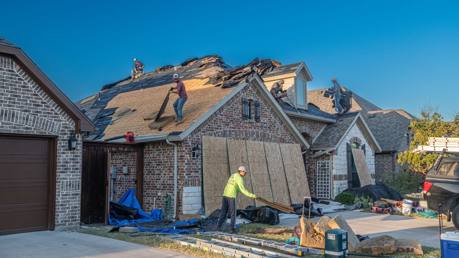 Roofing crew tearing off and replacing shingles on a brick house with a steep roof under a clear blue sky